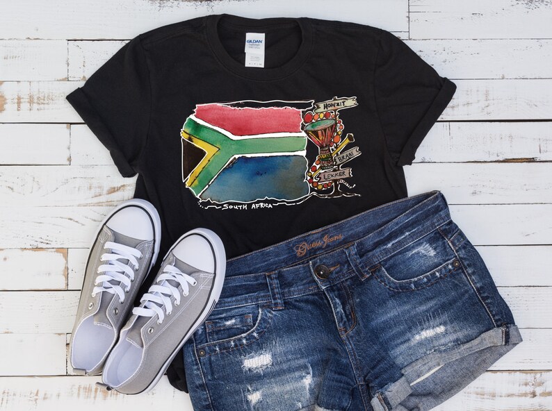 Puede incluir: Camiseta negra con un dise&ntilde;o de acuarela de la bandera sudafricana, con el texto "South Africa". La camiseta se combina con pantalones cortos de mezclilla y zapatillas de lona grises. El dise&ntilde;o incluye un tambor y otros instrumentos musicales.