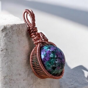 May include: A handcrafted pendant featuring a vibrant, multi-colored gemstone wrapped in copper wire. The gemstone displays a mix of purple, green, and blue hues. The copper wire is intricately woven, creating a cage-like design around the stone.