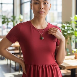 May include: A woman wearing a red t-shirt dress and a gold necklace with a round pendant. The dress has short sleeves and a gathered waist. The background includes windows, plants, and tables.