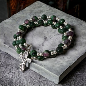 May include: A green and pink gemstone bracelet with a silver cross charm. The bracelet is made of small round beads and is double-stranded.