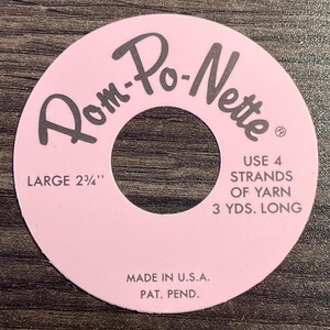 May include: A pink Rom-Po-Nette pom-pom maker with a central hole. The text reads "LARGE 2 3/4"" and "USE 4 STRANDS OF YARN 3 YDS. LONG." It also says "MADE IN U.S.A." and "PAT. PEND."