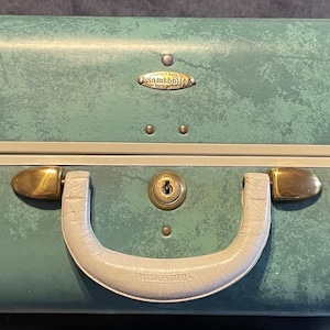 Puede incluir: Maleta vintage de color turquesa con herrajes dorados y asa y ribete color crema. La maleta presenta una superficie texturizada y una pequeña placa metálica. La maleta tiene dos cierres dorados y un ojo de cerradura.