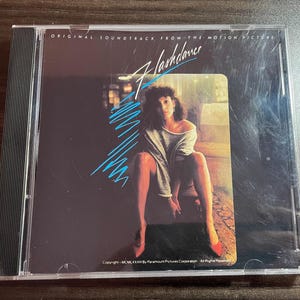 May include: A CD case featuring the "Flashdance" soundtrack. The cover art shows a woman in a gray top and red heels. The title "Flashdance" is in a stylized font with blue accents. The case is in a clear plastic sleeve.