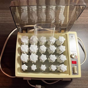 May include: Vintage General Electric hair curler set. The cream-colored base holds multiple white curlers of varying sizes. A clear plastic cover is open, revealing the curlers inside. The device has a power switch and indicator lights.