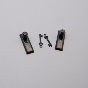 May include: Two black metal door knobs with keyholes and two matching keys. The knobs are rectangular with rounded edges and have a small, round knob on the top.