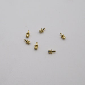 May include: Several small, gold-colored metal components are scattered on a white surface. Each component has a cylindrical body with a flared base and a small, protruding pin. The components appear to be new.
