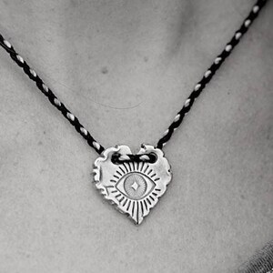 May include: A silver-toned heart-shaped pendant necklace with an eye design. The pendant hangs from a black and silver braided cord. The necklace is being held by a hand with multiple rings.