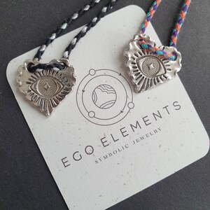 May include: Two silver-colored heart-shaped pendants with an eye design, suspended on braided cords. The pendants rest on a card with the text "EGO ELEMENTS SYMBOLIC JEWELRY". The cords are black and white, and multicolored.