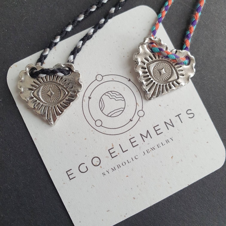 May include: Two silver-colored heart-shaped pendants with an eye design, suspended on braided cords. The pendants rest on a card with the text "EGO ELEMENTS SYMBOLIC JEWELRY". The cords are black and white, and multicolored.