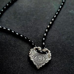May include: A silver-toned heart pendant necklace with an eye design. The pendant is suspended from a black cord with white dots. The eye design features a central star and radiating lines, set against a dark background.