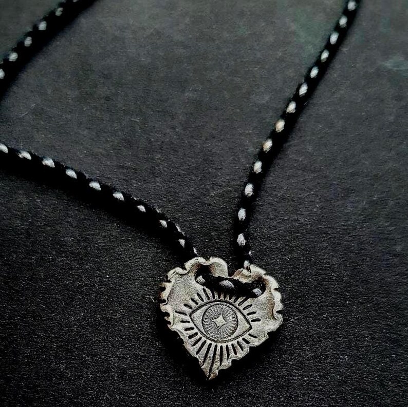 May include: A silver-toned heart pendant necklace with an eye design. The pendant is suspended from a black cord with white dots. The eye design features a central star and radiating lines, set against a dark background.