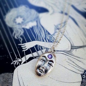 May include: Silver necklace with a face pendant featuring a purple gemstone in the third eye. The pendant is oval shaped and has a detailed face with closed eyes.
