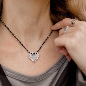 May include: A silver-colored pendant necklace with a heart-shaped charm. The charm features an eye design and is suspended from a black and white braided cord. The necklace is worn against a gray shirt.