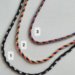 May include: Three braided cord necklaces in black, multicolored, and purple. The black cord has silver accents. The multicolored cord features brown, pink, yellow, and blue. The purple cord has orange, teal, and gray accents. The cords are displayed on a light gray surface.