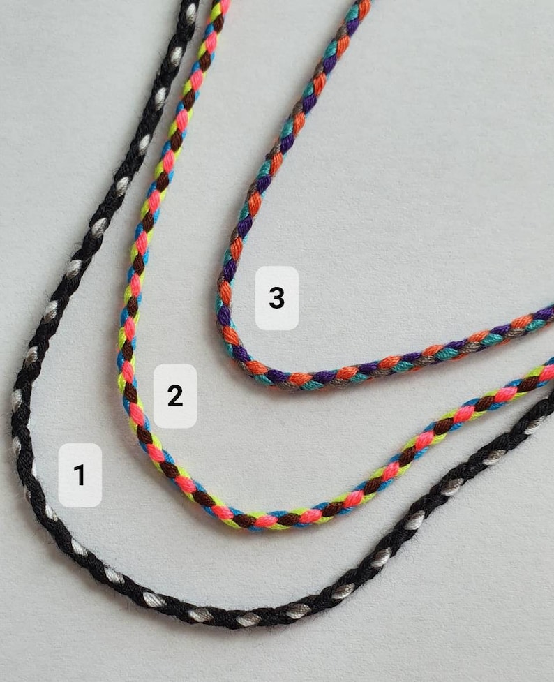 May include: Three braided cord necklaces in black, multicolored, and purple. The black cord has silver accents. The multicolored cord features brown, pink, yellow, and blue. The purple cord has orange, teal, and gray accents. The cords are displayed on a light gray surface.