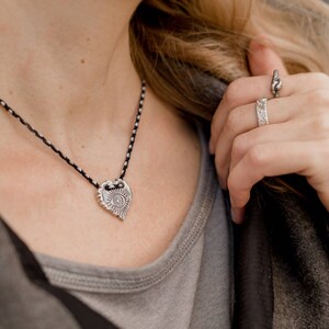 May include: Silver heart-shaped pendant necklace with a black and white cord. The pendant features an eye design with sun rays. A silver ring with a textured design is also visible.