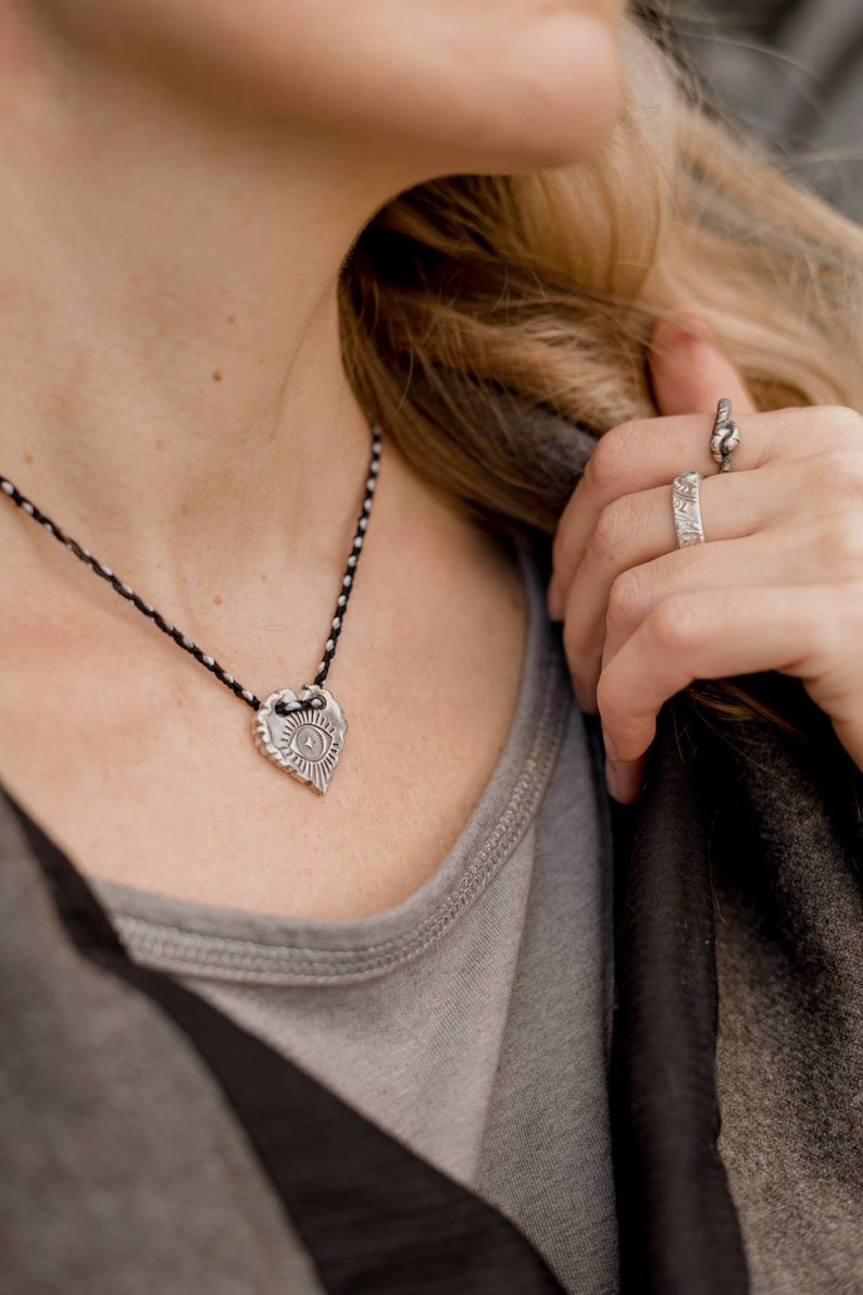May include: Silver heart-shaped pendant necklace with a black and white cord. The pendant features an eye design with sun rays. A silver ring with a textured design is also visible.