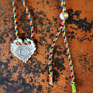 May include: A silver-colored pendant necklace with a heart-shaped design featuring an eye symbol. The necklace has a colorful braided cord in shades of pink, yellow, blue, and black. The necklace is displayed on a brown, textured surface.