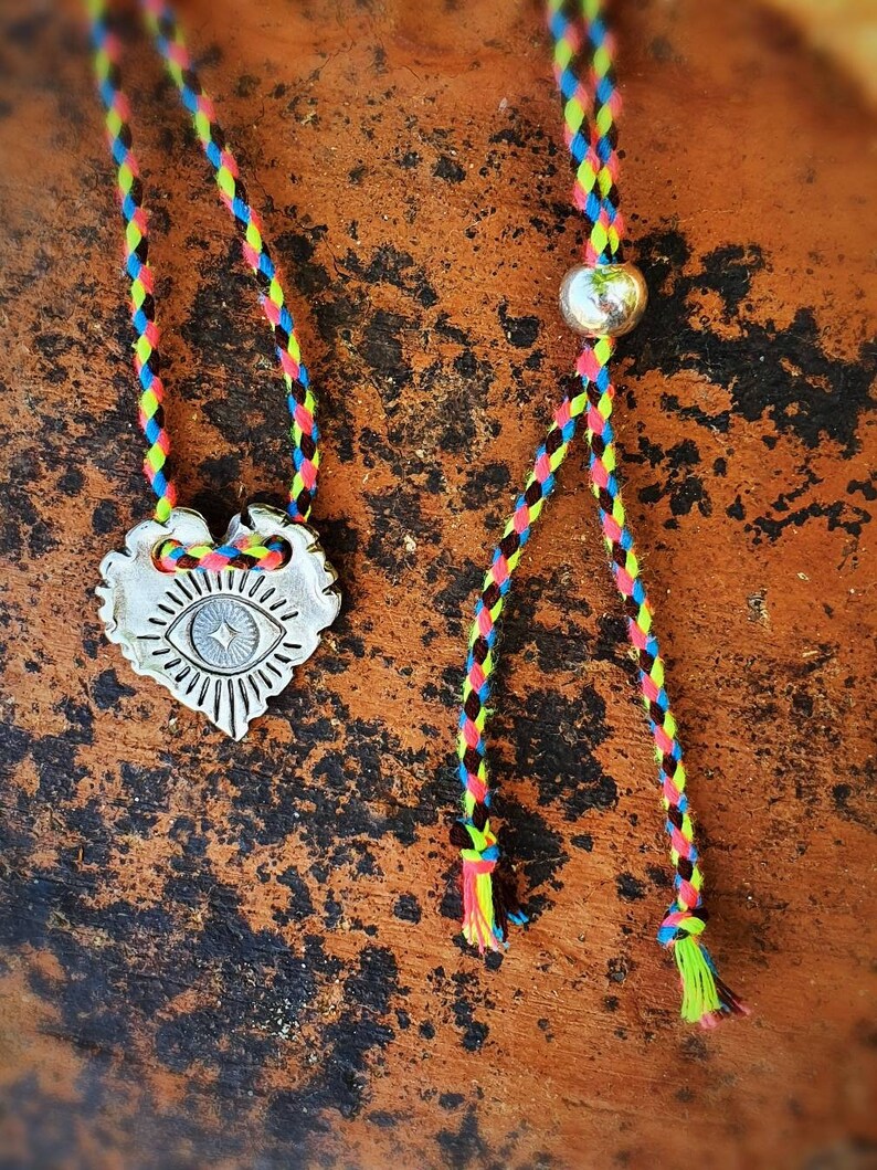 May include: A silver-colored pendant necklace with a heart-shaped design featuring an eye symbol. The necklace has a colorful braided cord in shades of pink, yellow, blue, and black. The necklace is displayed on a brown, textured surface.