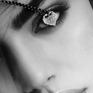 May include: Black and white image of a silver heart-shaped pendant with an eye design. The pendant hangs from a black and white cord. The image is a close-up of a person's eye and face.