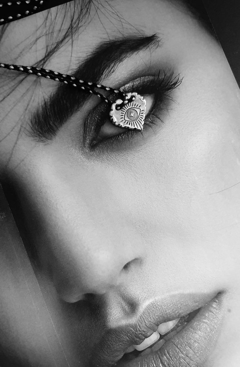 May include: Black and white image of a silver heart-shaped pendant with an eye design. The pendant hangs from a black and white cord. The image is a close-up of a person's eye and face.