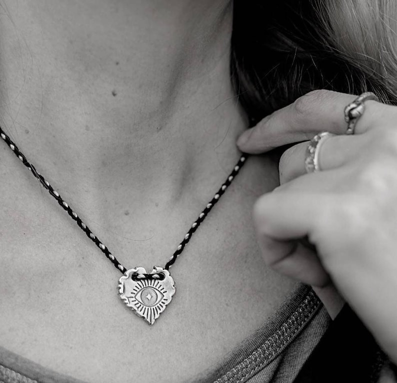 May include: A silver-toned heart-shaped pendant necklace with an eye design. The pendant hangs from a black and silver braided cord. The necklace is being held by a hand with multiple rings.