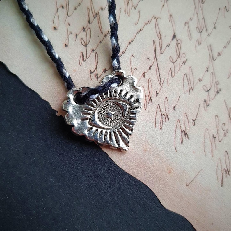 May include: A silver heart-shaped pendant with an eye design, suspended from a black and white braided cord. The pendant has a textured surface and is set against a backdrop of aged paper with handwritten script.