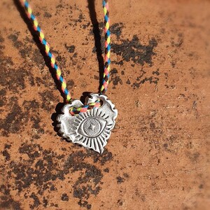 May include: A silver-colored heart pendant necklace with an eye design. The pendant is suspended from a colorful braided cord in shades of blue, yellow, red, and brown. The necklace is displayed on a textured brown surface.