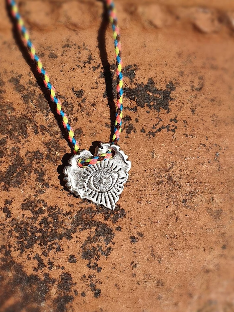 May include: A silver-colored heart pendant necklace with an eye design. The pendant is suspended from a colorful braided cord in shades of blue, yellow, red, and brown. The necklace is displayed on a textured brown surface.
