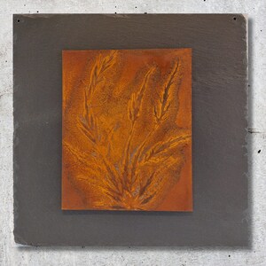 Könnte beinhalten: Ein quadratisches Stück Schiefer mit einem quadratischen Einsatz aus rostigem orangefarbenem Metall in der Mitte. Das Metallquadrat zeigt ein Design einer stilisierten Pflanze mit mehreren Stielen und Blättern.