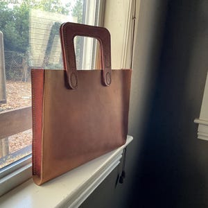 May include: A brown leather tote bag with a rectangular shape and a matching handle. The bag has a contrasting pink side panel and is sitting on a white windowsill. The handle is stitched to the bag.