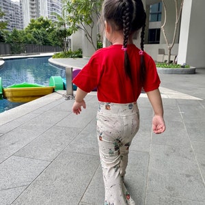 May include: A young child wearing a red t-shirt and beige pants with a cactus print. The pants have a flared leg design. The child is also wearing pink sandals and has their hair in two braids.