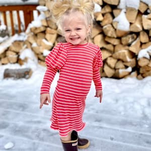 May include: A young girl wearing a pink and white striped dress with long sleeves and puffed shoulders. She is standing in the snow and wearing brown rain boots.