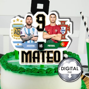 May include: A cake topper featuring Lionel Messi and Cristiano Ronaldo, with the text "Argentina vs Portugal" and "Mateo". The topper is decorated with the flags of Argentina and Portugal.