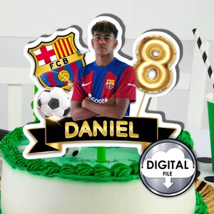 May include: A cake topper featuring a young boy wearing a red and blue soccer jersey with the FC Barcelona logo. The topper also includes a gold number 8 balloon, a soccer ball, and a black banner with the name "DANIEL" in gold letters.