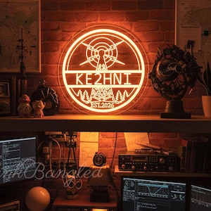 Könnte beinhalten: Ein leuchtendes orangefarbenes Neon-Schild mit dem Text "KE2HNI" und "EST.2026" ist an einer Backsteinmauer angebracht. Das Schild zeigt ein Funkmast-Design in einem kreisförmigen Rahmen. Die Szene umfasst einen Schreibtisch mit Computermonitoren und Funkanlagen.