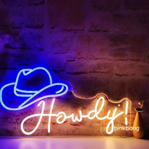 以下が含まれることがあります： 白いネオンサインに、筆記体で「Howdy!」の文字が書かれています。青いネオンの カウボーイハットが「Howdy!」の文字の上にあります。サインはレンガの壁に取り付けられています。