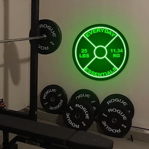 Może przedstawiać: Zielony neonowy znak z napisem "Everyday Essentials" i wzorem talerza do ćwiczeń. Znak pokazuje 25 funtów i 11,34 kg. Znak wisi na ścianie w domowej siłowni z ławką do ćwiczeń i talerzami do ćwiczeń.