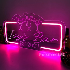 Puede incluir: Un letrero de neón rosa para "Ivy's Bar" con ilustraciones de cócteles y el texto "EST 2023". El letrero tiene forma rectangular con una parte superior curva y un borde rosa brillante. Se ven botellas a ambos lados, acentuando el tema del bar.