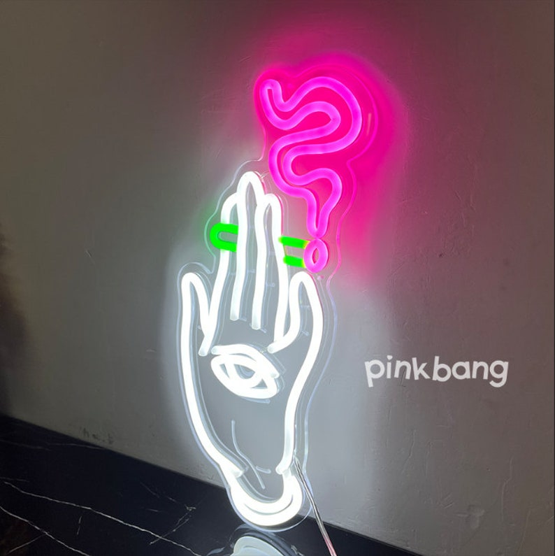 May include: White neon sign of a hand holding a cigarette with pink neon smoke rising above it. The sign says "pinkbang" in white text.