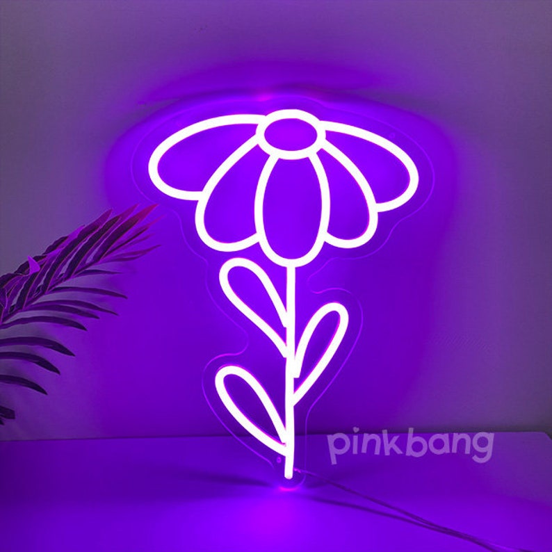 May include: A white neon sign shaped like a flower with a purple glow. The sign is on a purple wall with a green plant in the background. The text "pinkbang" is visible in the bottom right corner of the image.