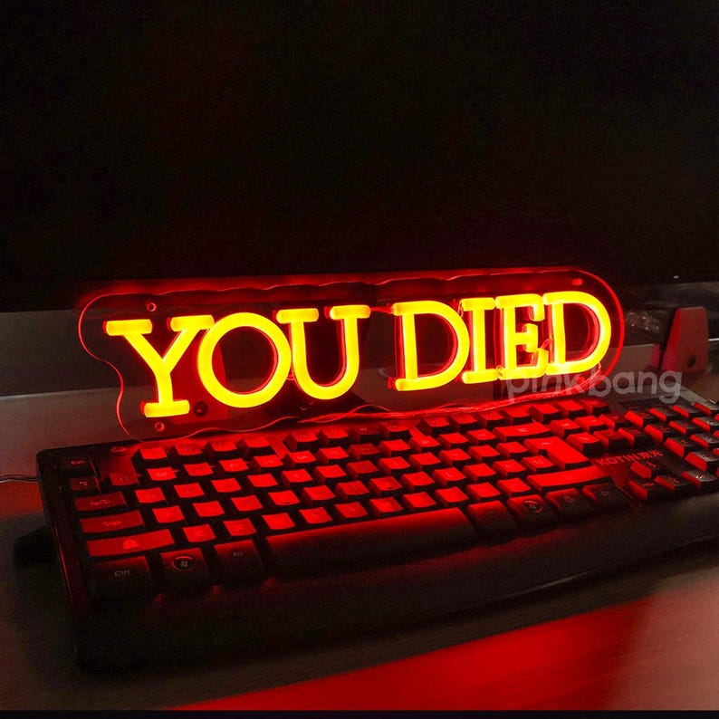 May include: A red neon sign with the words "YOU DIED" in yellow letters. The sign is sitting on top of a black keyboard.