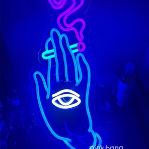 May include: A blue neon sign depicting a hand holding a cigarette with smoke rising above it. The hand has an eye in the palm. The sign has the text "pinkbang" at the bottom.
