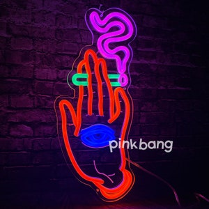 May include: Neon sign depicting a hand holding a cigarette with smoke rising above it. The hand has an eye in the palm. The sign is lit in red, blue, green, and purple neon colors. The text "pinkbang" is visible in the lower right corner.