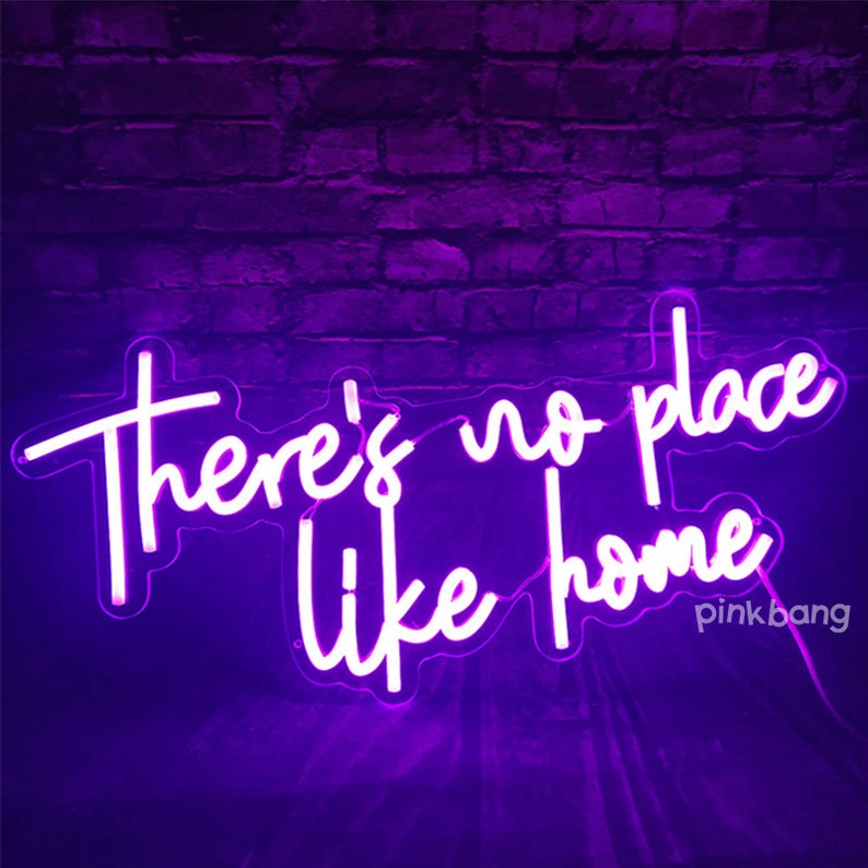 May include: Purple neon sign with white lettering that reads "There's no place like home". The sign is on a brick wall background.