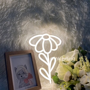 May include: White neon sign shaped like a daisy flower with a stem and leaves. The sign is lit up and is sitting on a white fluffy surface.