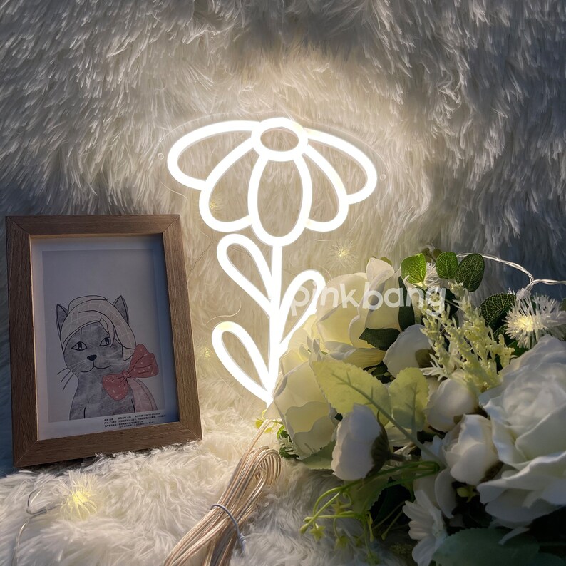 May include: White neon sign shaped like a daisy flower with a stem and leaves. The sign is lit up and is sitting on a white fluffy surface with a bouquet of white flowers and a framed picture of a cat.