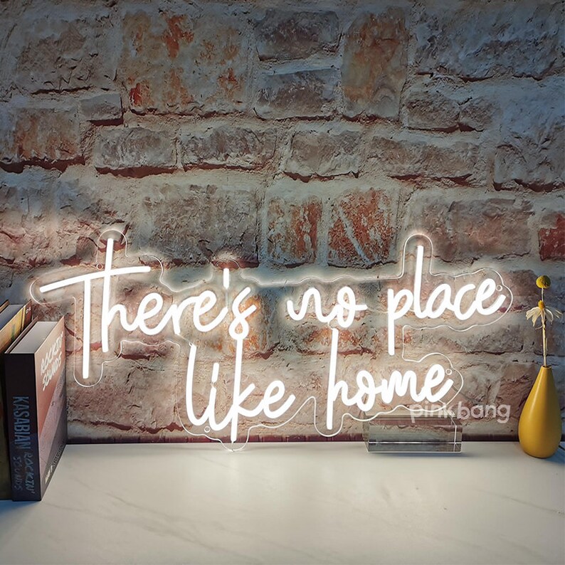 May include: White neon sign with the text "There's no place like home". The sign is mounted on a clear acrylic base and is sitting on a wooden surface.