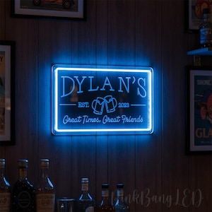 Puede incluir: Un letrero de neón azul con el nombre "DYLAN'S" y el texto "Great Times, Great Friends". El letrero incluye el año "2023" e imágenes de jarras de cerveza. El letrero es rectangular y está montado en una pared de madera.