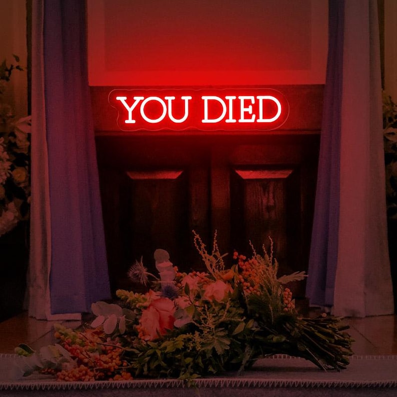 May include: A red neon sign with the words "YOU DIED" in white lettering. The sign is mounted on a wooden surface above a bouquet of flowers.